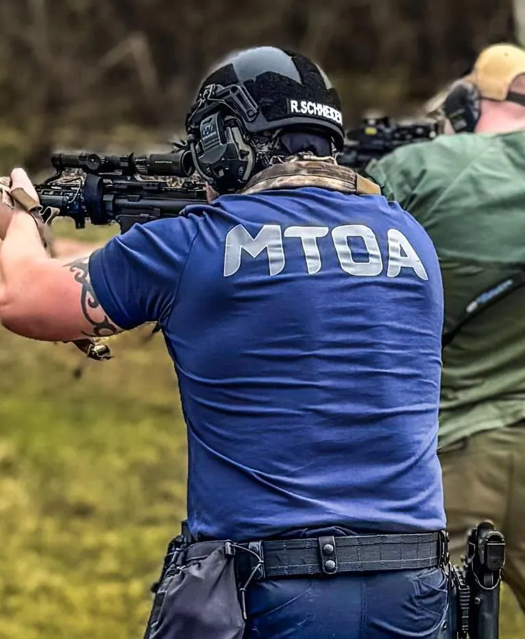 Two men facing away from the camera with rifles at a gun range. One is wearing a blue shirt with large 'MTOA' text on the back.